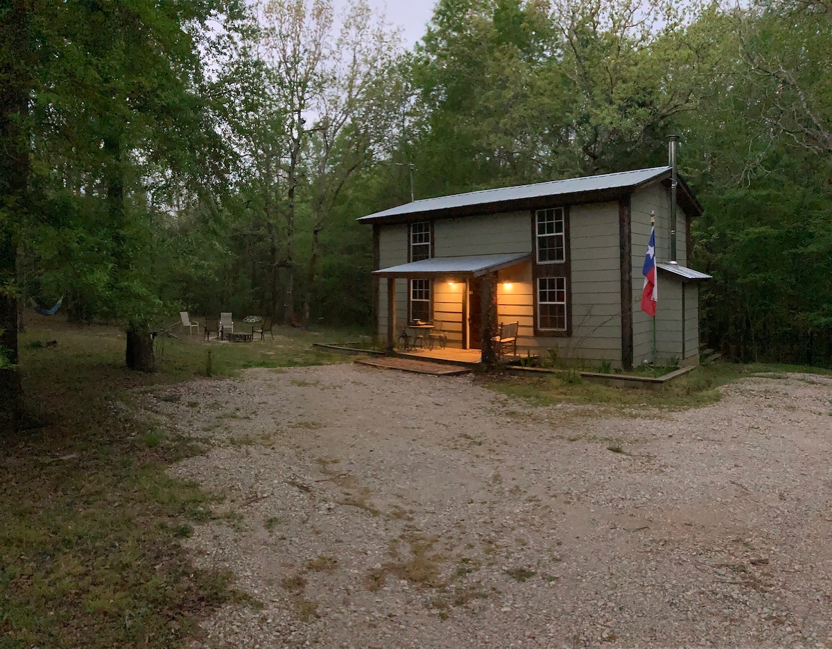 A quiet cabin framed by greenery is shown during twilight. The inviting porch, with light glowing from the windows, is complemented by a gravel drive. Several outdoor chairs are arranged nearby, creating a peaceful setting in a natural landscape.