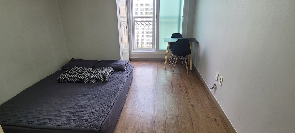 A simple sleeping area features a spacious bed with a patterned pillow placed at the head. A small dining table with chairs is situated nearby, and natural light can be seen streaming through a large window, enhancing the minimalist decor.