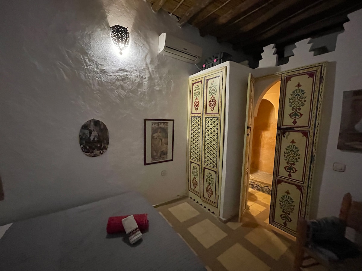 A spacious room offers a blend of traditional décor with a colorful wardrobe and textured walls. A bedside table beside a restful sleeping area is adorned with a red towel. An arched doorway leads to an additional space, complemented by soft ambient lighting.