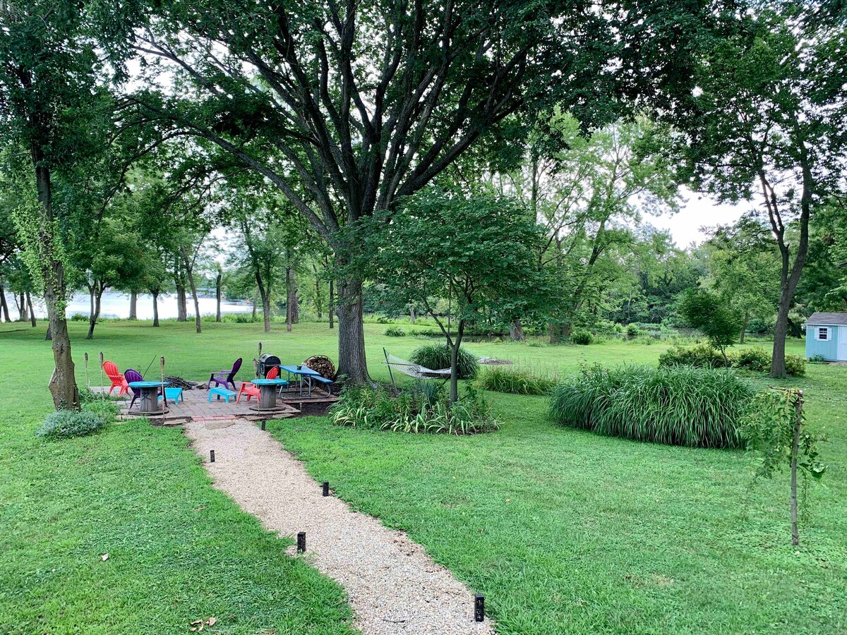 A scenic outdoor space features a winding gravel pathway leading to a vibrant seating area with colorful chairs around a fire pit. A hammock hangs nearby, surrounded by lush greenery and trees. The lake view is visible in the background, enhancing the tranquil atmosphere.