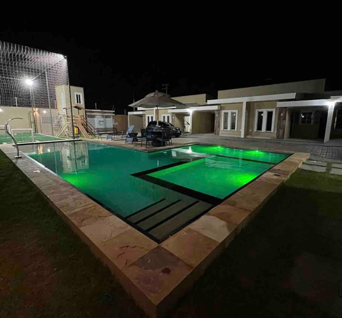 A spacious outdoor area is highlighted by a large pool, illuminated in green light against the night sky. Surrounding the pool, a lawn is visible alongside modern seating arrangements. The setting showcases a relaxed atmosphere, with the pool area complemented by well-defined edges and a clean design.