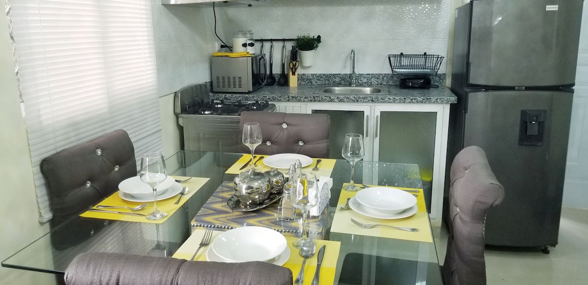 A modern kitchen area features a glass dining table set for four, complete with plates and wine glasses. The kitchen is equipped with a refrigerator, stove, and sink, while overhead cabinets provide storage. Natural light enters through a nearby window, brightening the space.