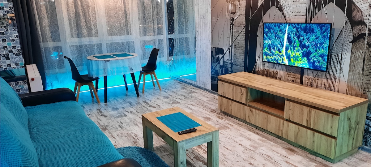 A modern living area is showcased with a sleek television on a wooden media console. A round dining table with two black chairs is positioned nearby. Soft lighting enhances the atmosphere, complemented by a blue accent wall and light wooden flooring.