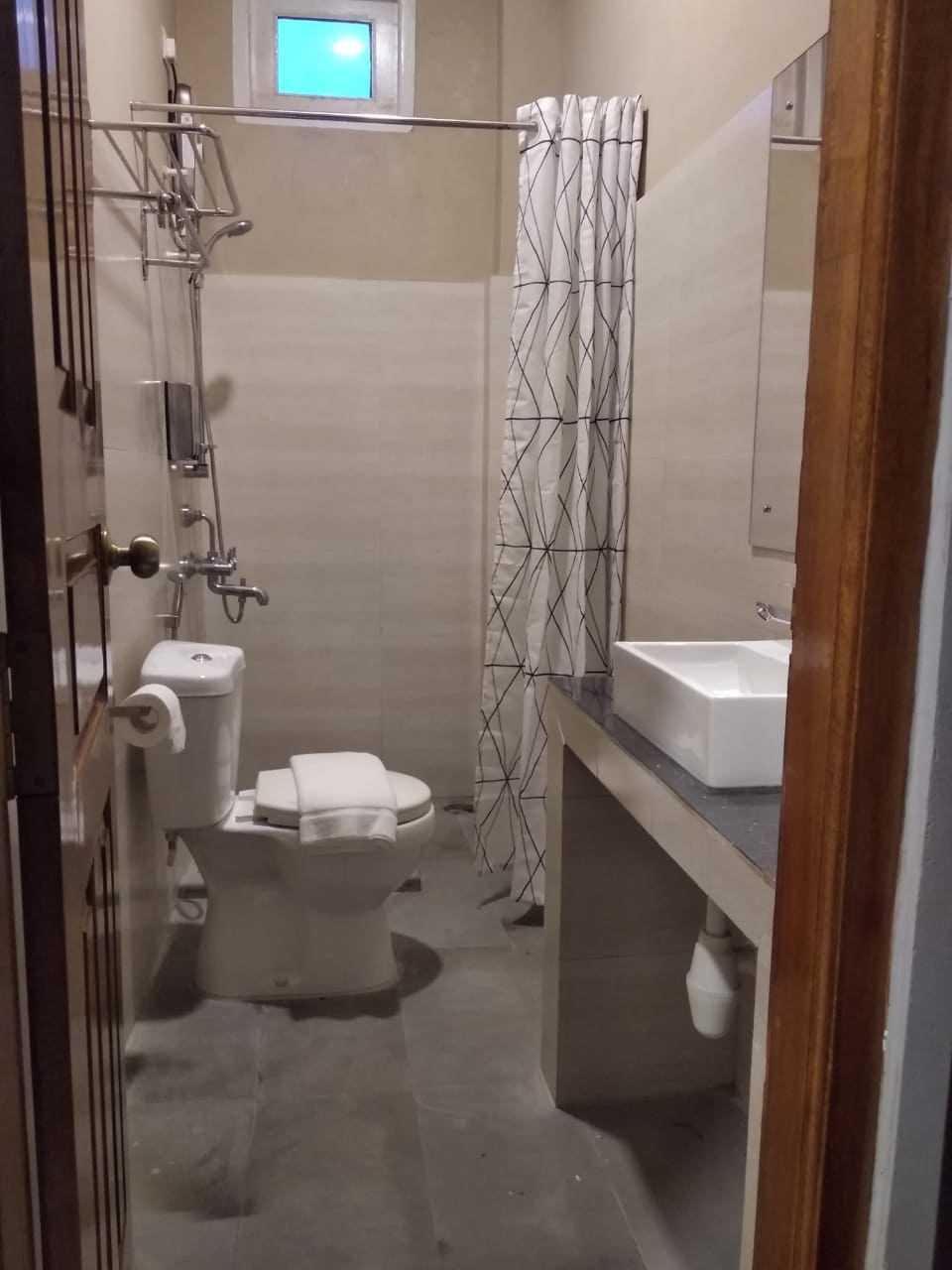 A bathroom space features a white toilet and a wall-mounted sink atop a dark countertop. A shower area is enclosed by a patterned curtain, and a small window above allows natural light to enter. Neutral walls and gray flooring create a cohesive design.