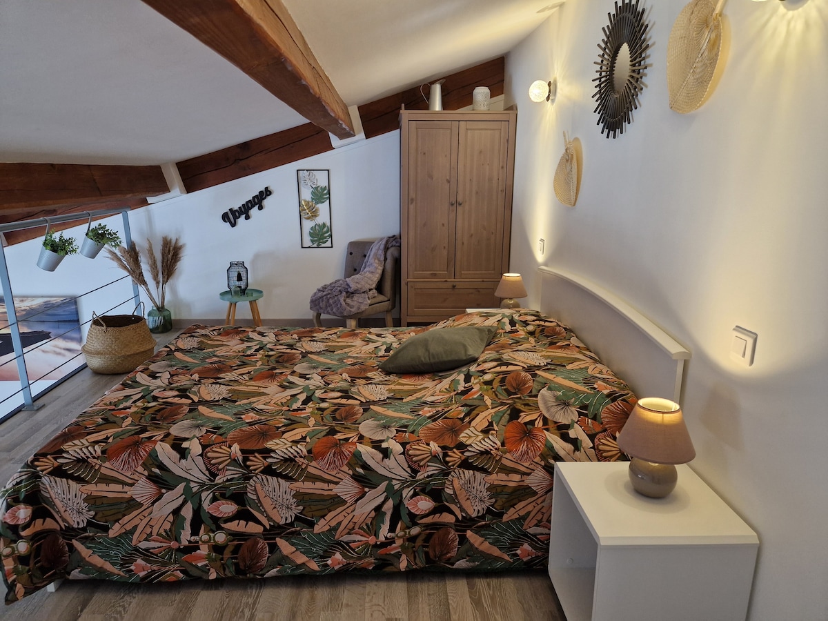 The sleeping area features a spacious bed dressed in a nature-inspired patterned coverlet. A wooden wardrobe stands against the wall, complemented by decorative wall accents. Soft lighting is provided by bedside lamps, enhancing the relaxed atmosphere. A chair with a cozy throw is also present.