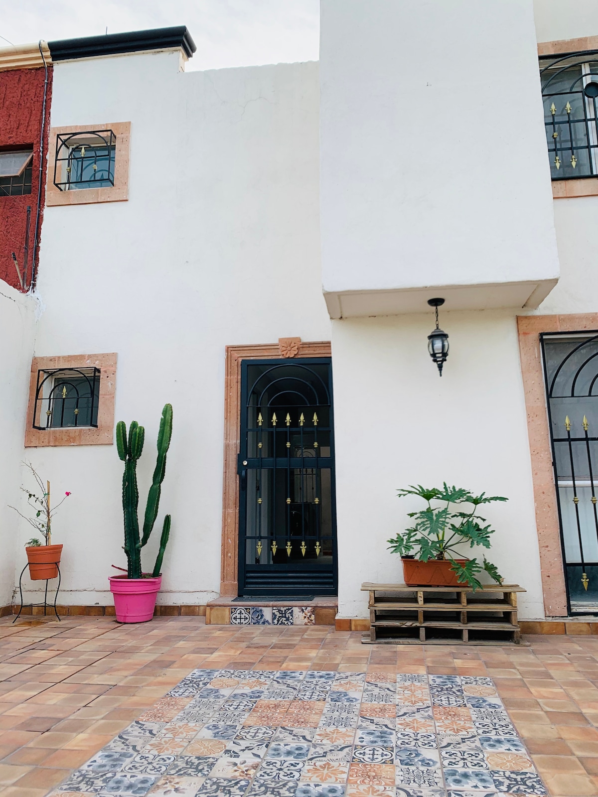 The entrance of the property is highlighted by a decorative door framed by elegant ironwork. Potted plants, including a tall cactus and greenery, add a touch of nature. The tiled pathway features a mix of patterns, offering a welcoming approach to the home.