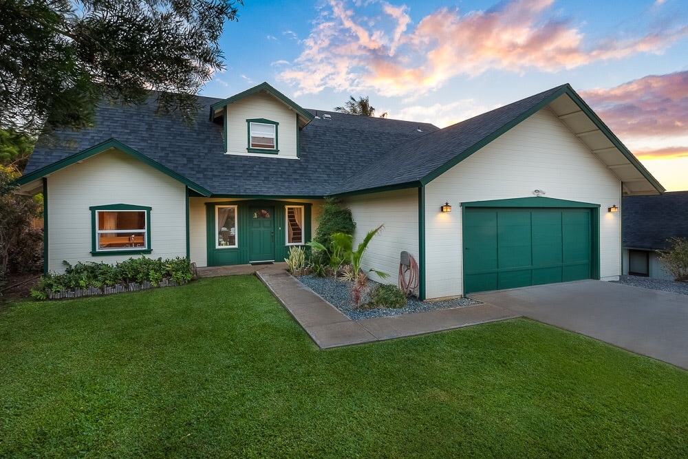 A two-story house is featured, showcasing a spacious front lawn with well-maintained greenery. The exterior is painted in light colors with green accents, including a double garage. Sunlight illuminates the home, which is set against a sky with vibrant sunset hues.