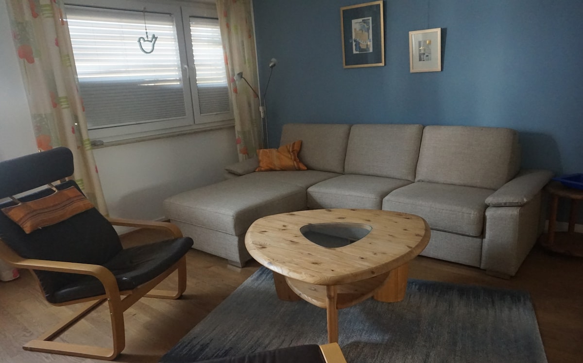 A comfortable living area features a spacious sectional sofa with a light grey fabric and a unique wooden coffee table with a cutout design. A dark upholstered chair complements the space, while decorative curtains add subtle colors to the room.