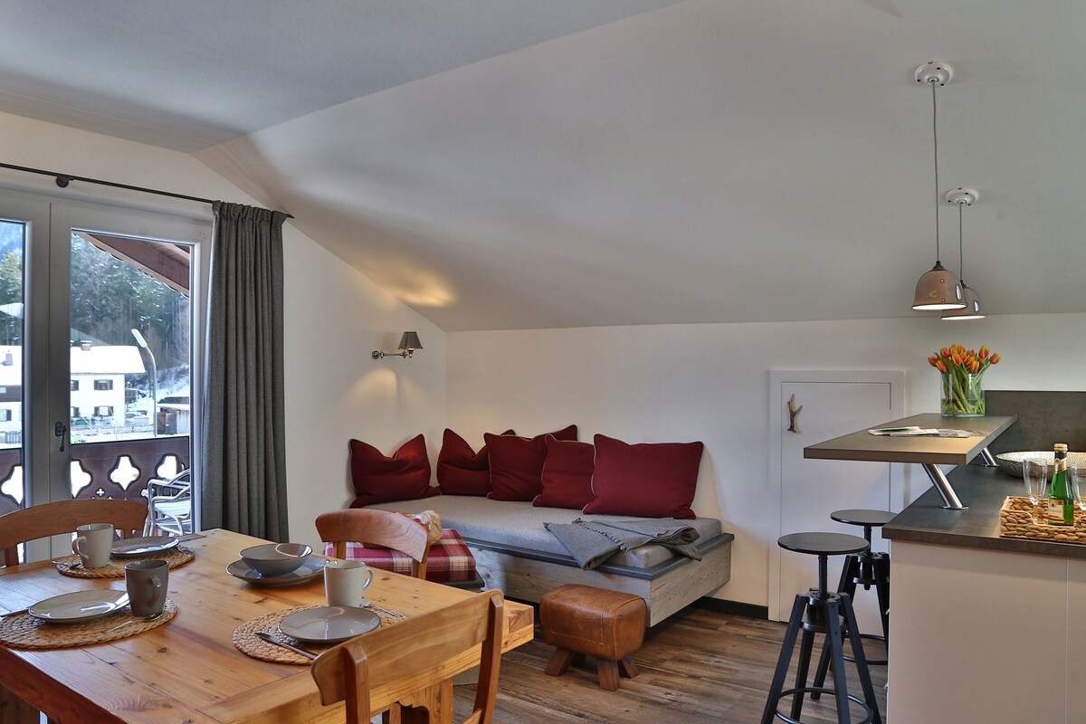 A bright dining area features a wooden table set for two, complemented by simple gray dishes. A corner sofa adorned with red cushions adds comfort, while natural light streams in through the large glass doors leading to the balcony, enhancing the inviting atmosphere.