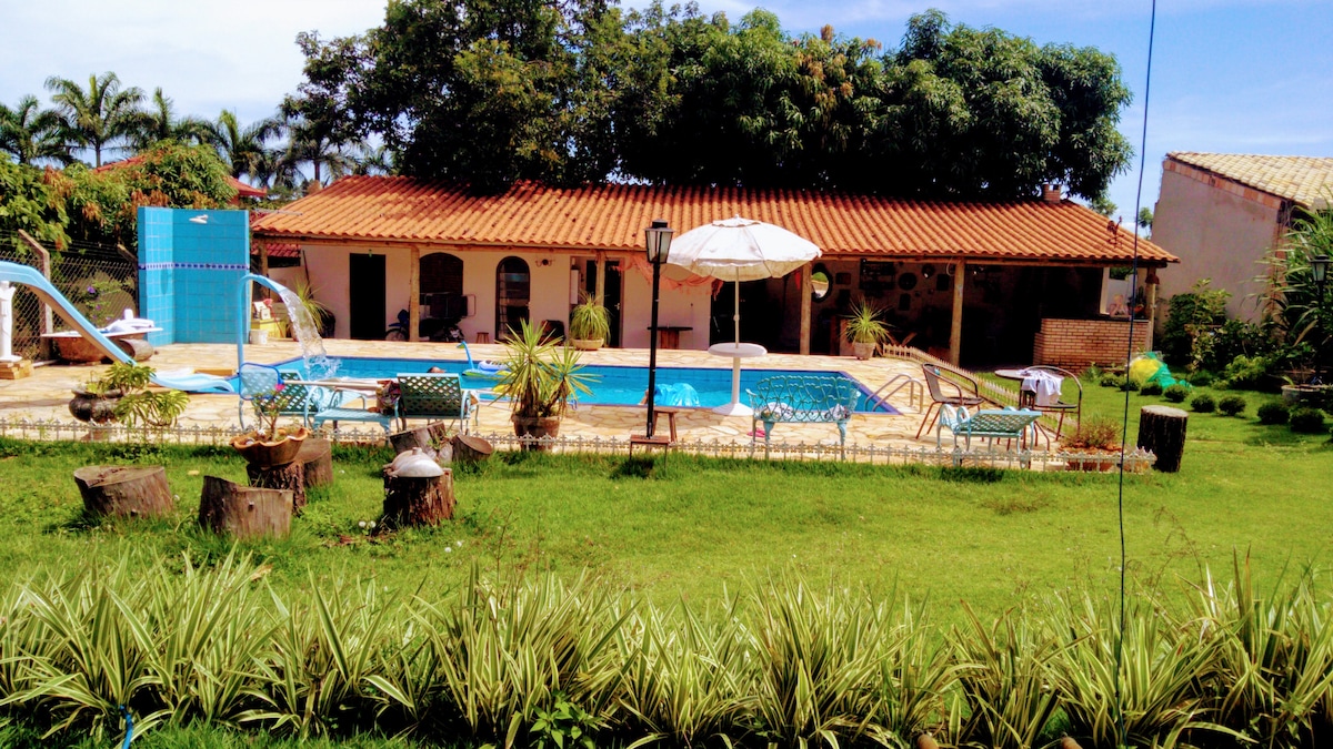 A spacious outdoor area features a private pool surrounded by sun loungers and umbrellas. Lush green grass and ornamental plants frame the scene, while multiple trees provide shade. The charming house with a red-tiled roof stands in the background, enhancing the natural setting.