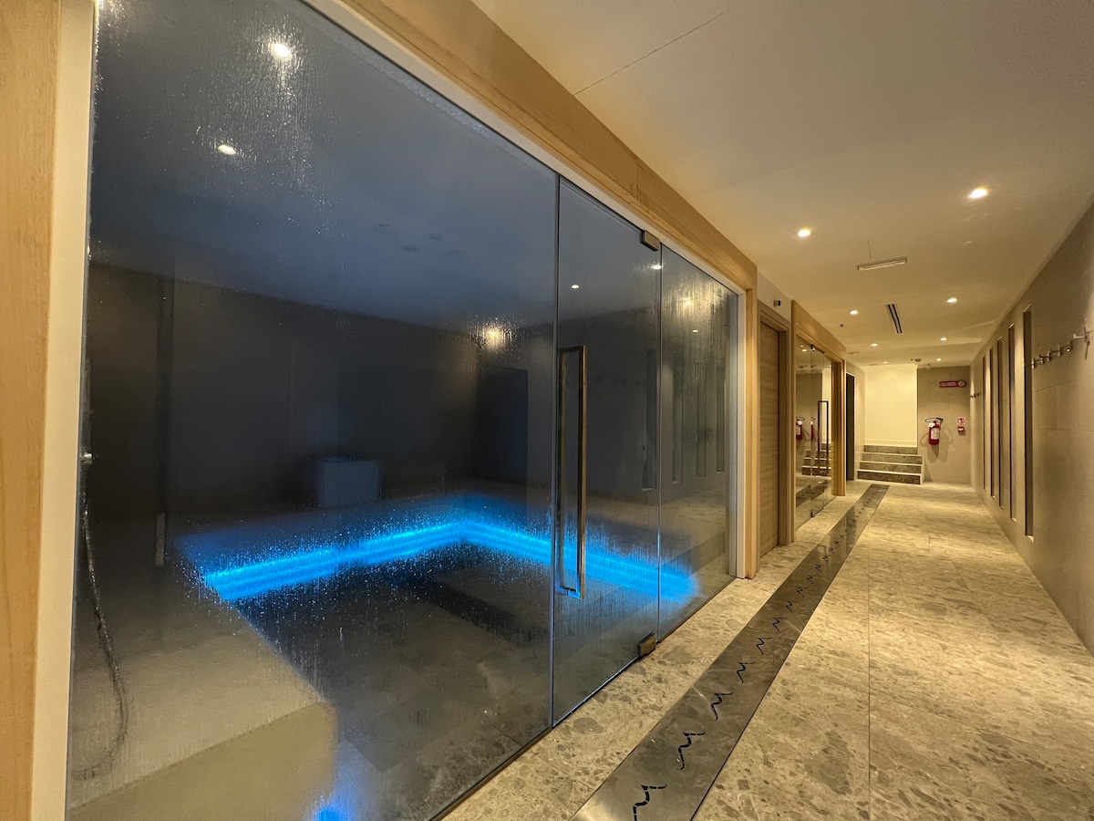 An elegant spa area is featured, illuminated by soft blue lighting. A large glass door separates the spa from the hallway, showcasing a serene hydro massage pool. The surrounding walls are adorned with smooth surfaces, and the floor is laid with polished stone.