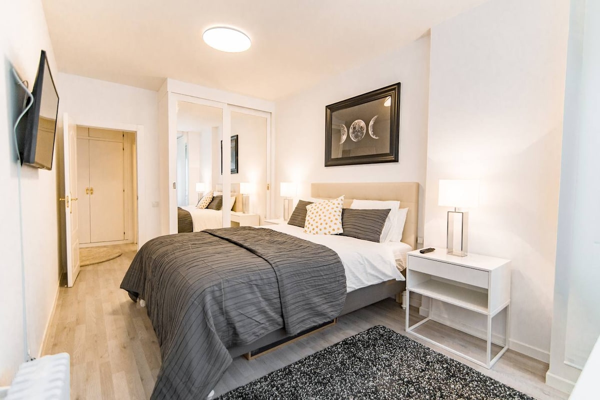 The bedroom features a spacious bed along with a striking piece of wall art. A flat-screen TV is mounted on the wall, complemented by a modern bedside table and a lamp. Bright light is provided by a ceiling fixture, creating a serene atmosphere.