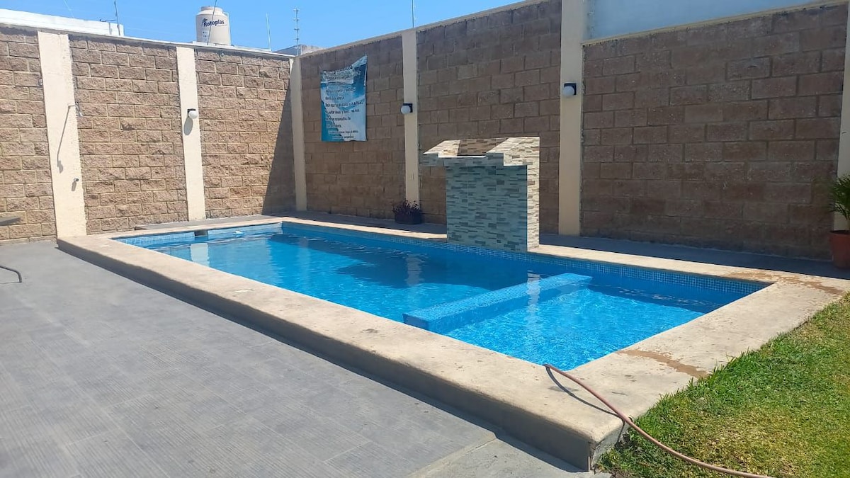 An inviting outdoor space features a large swimming pool surrounded by smooth concrete and grass areas. A stone structure with a built-in grill is positioned next to the pool, enhancing the functionality of the space. The walls are made of textured stone, adding a rustic touch.