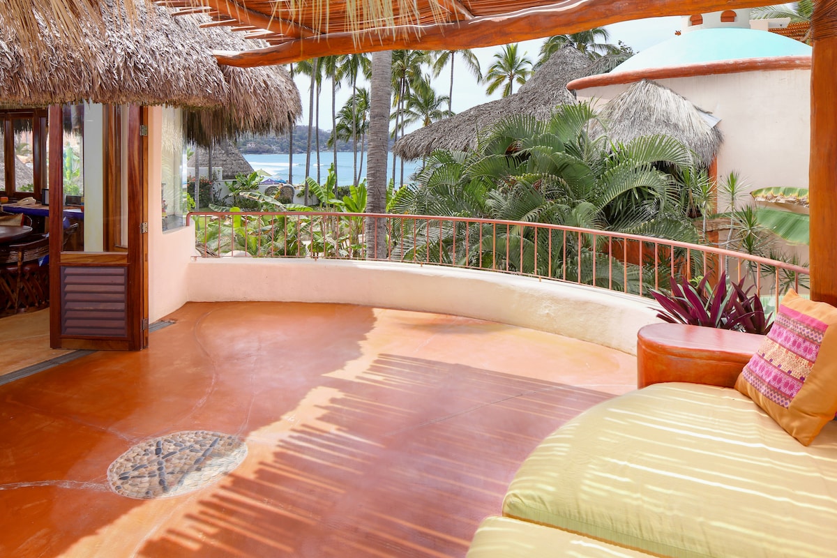 A circular terrace features a thatched roof and overlooks lush tropical landscaping toward the ocean. A comfortable, oversized built-in couch with bright cushions provides a relaxing seating area. The warm-toned flooring complements the natural surroundings, creating an inviting outdoor space.