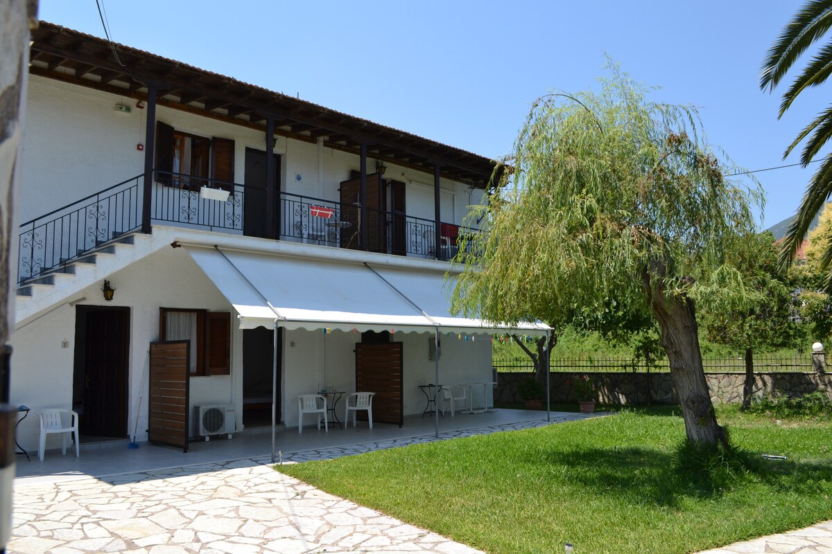 An inviting outdoor area is featured, showcasing a shaded patio with several white chairs and tables. The two-story building has a balcony providing access to upstairs rooms. Lush green grass surrounds the space, complemented by a large tree and distant hills visible in the background.