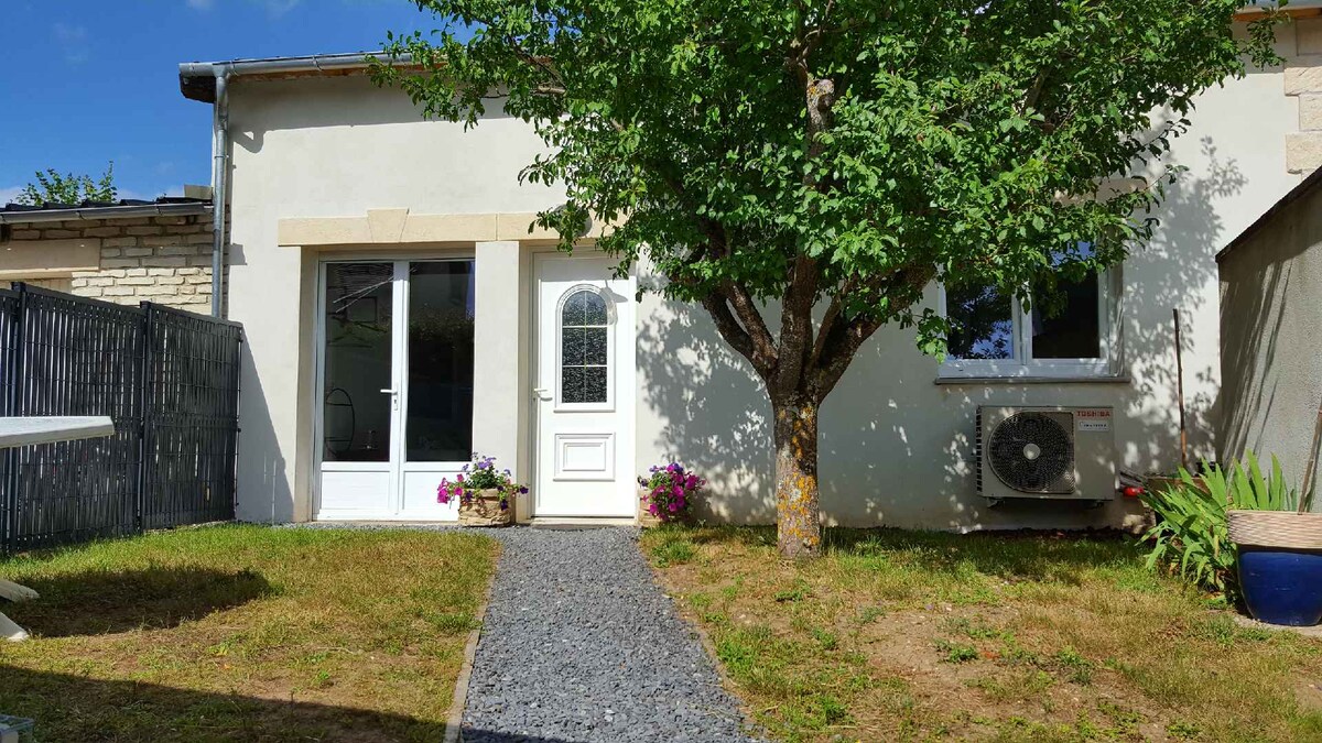 The entrance to the accommodation is framed by a grassy yard featuring a small tree and colorful potted flowers. A pathway of gravel leads to the white door, complemented by large windows that allow natural light. An air conditioning unit is visible on the side wall.