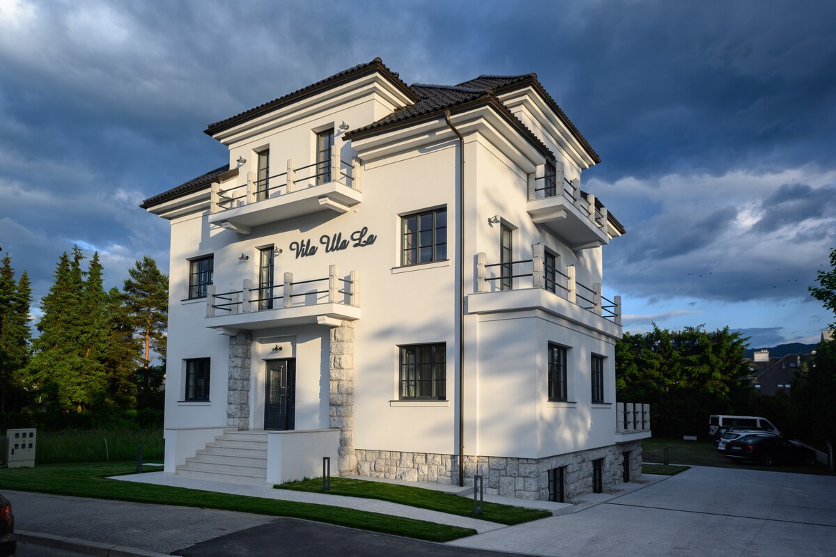 The villa is depicted with a modern architectural design, featuring a white exterior with gray stone accents. Multiple balconies are visible on the upper floors, along with large windows allowing for ample natural light. A well-maintained pathway leads to the entrance.