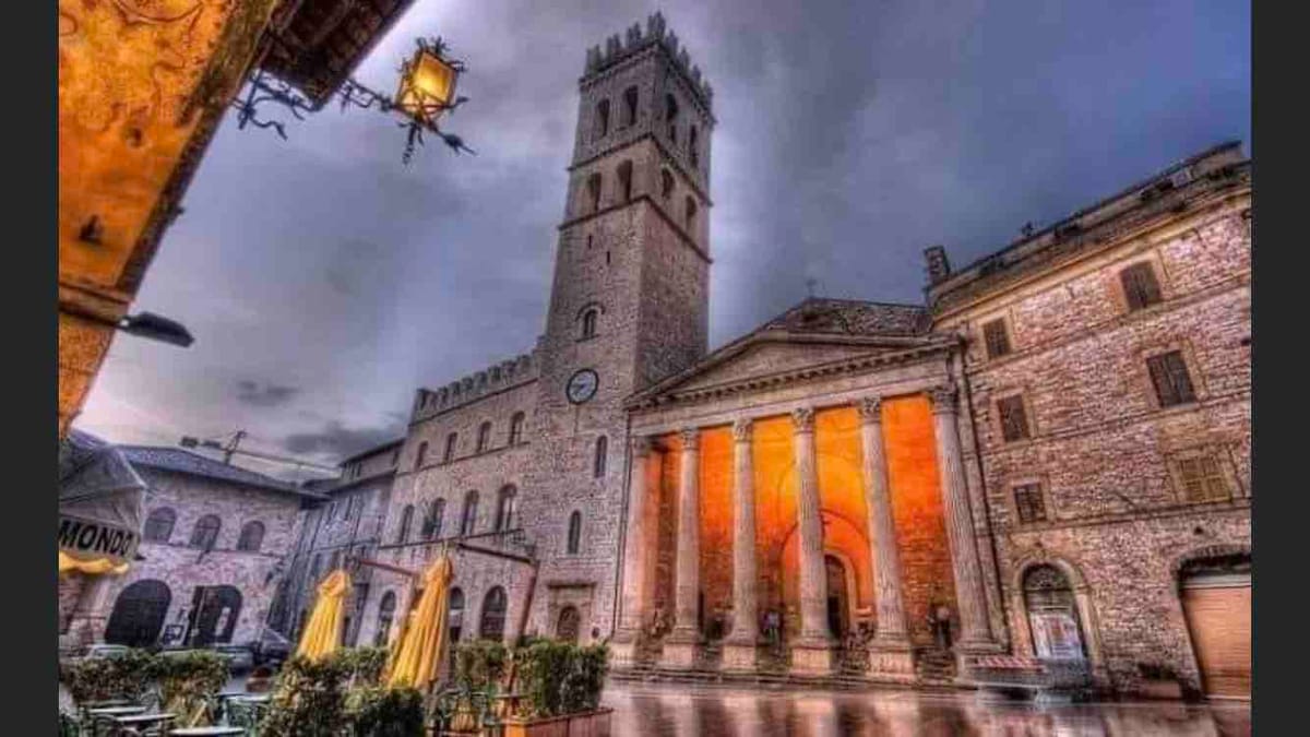 The image depicts a historic building with a tall clock tower adjacent to an elegant structure featuring multiple columns. The scene is set against a cloudy sky, and cafe tables with yellow umbrellas are visible in the foreground, reflecting a tranquil atmosphere.