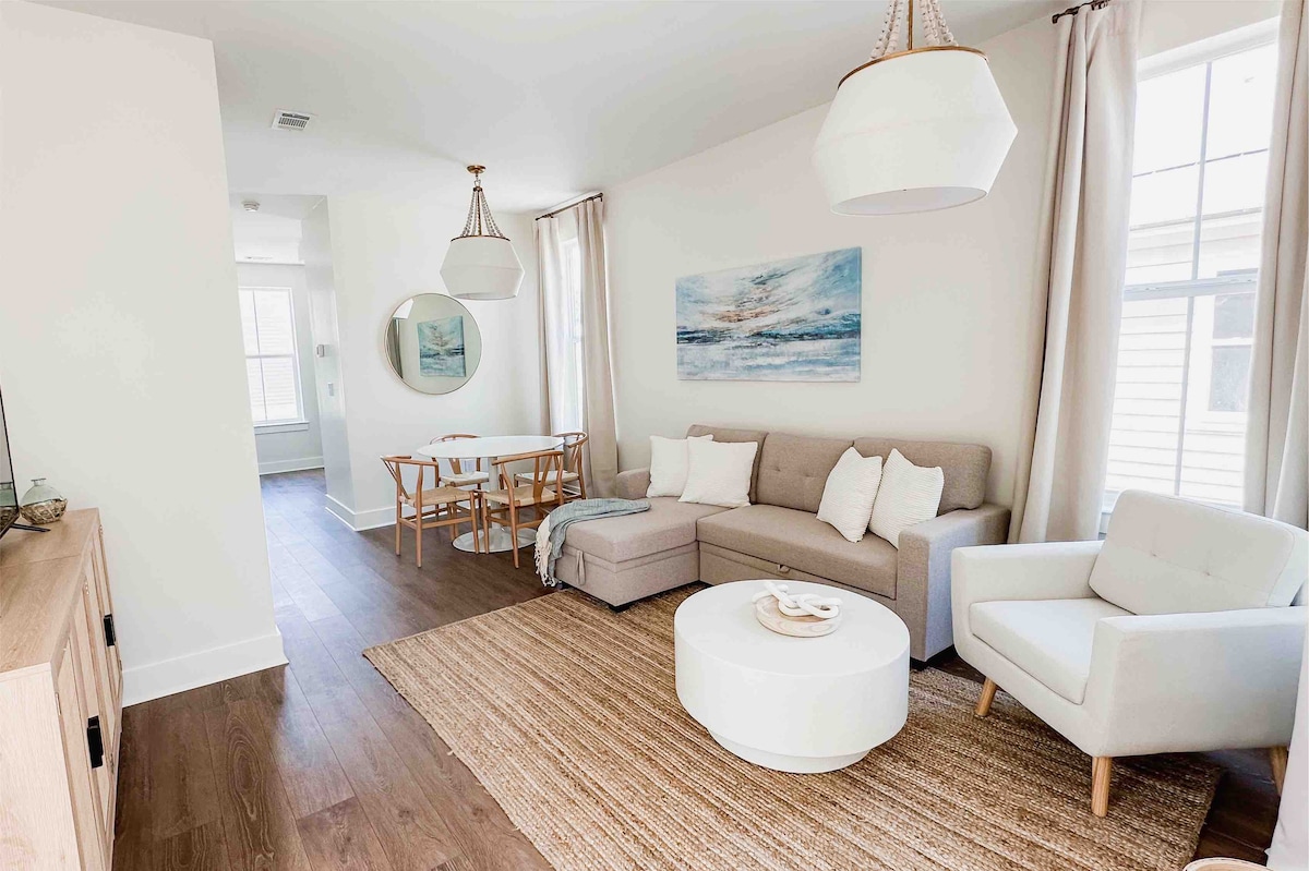 A bright living area features a light-colored sectional sofa with decorative cushions, accompanied by a sleek white coffee table. A dining table with wooden chairs is visible in the background, while large windows allow natural light to enhance the space, complemented by neutral curtains.
