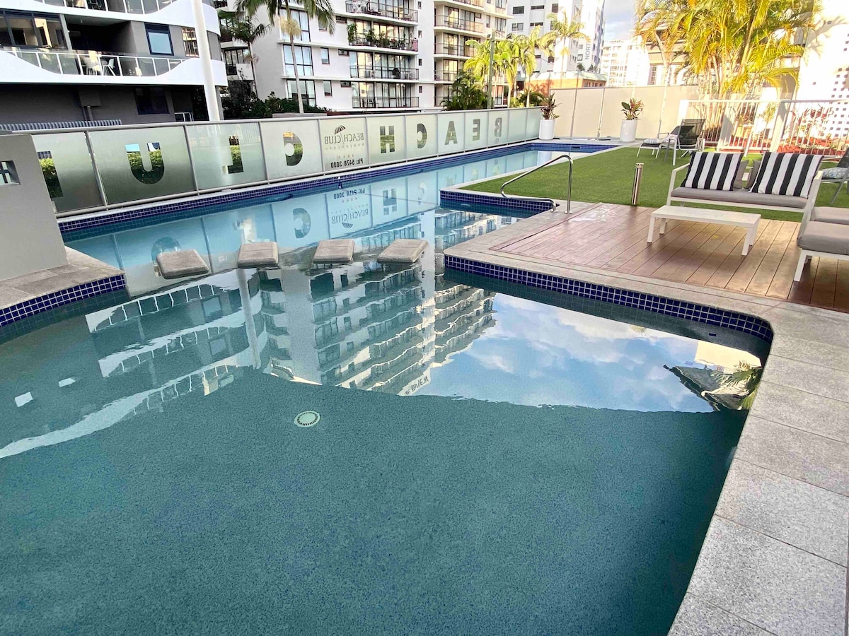 The resort's pool area features a spacious swimming pool with clear blue water reflecting the sky. Comfortable lounge chairs are positioned around the pool, alongside well-maintained grass areas and elegant outdoor seating. The surrounding buildings provide a modern backdrop, creating a serene atmosphere.