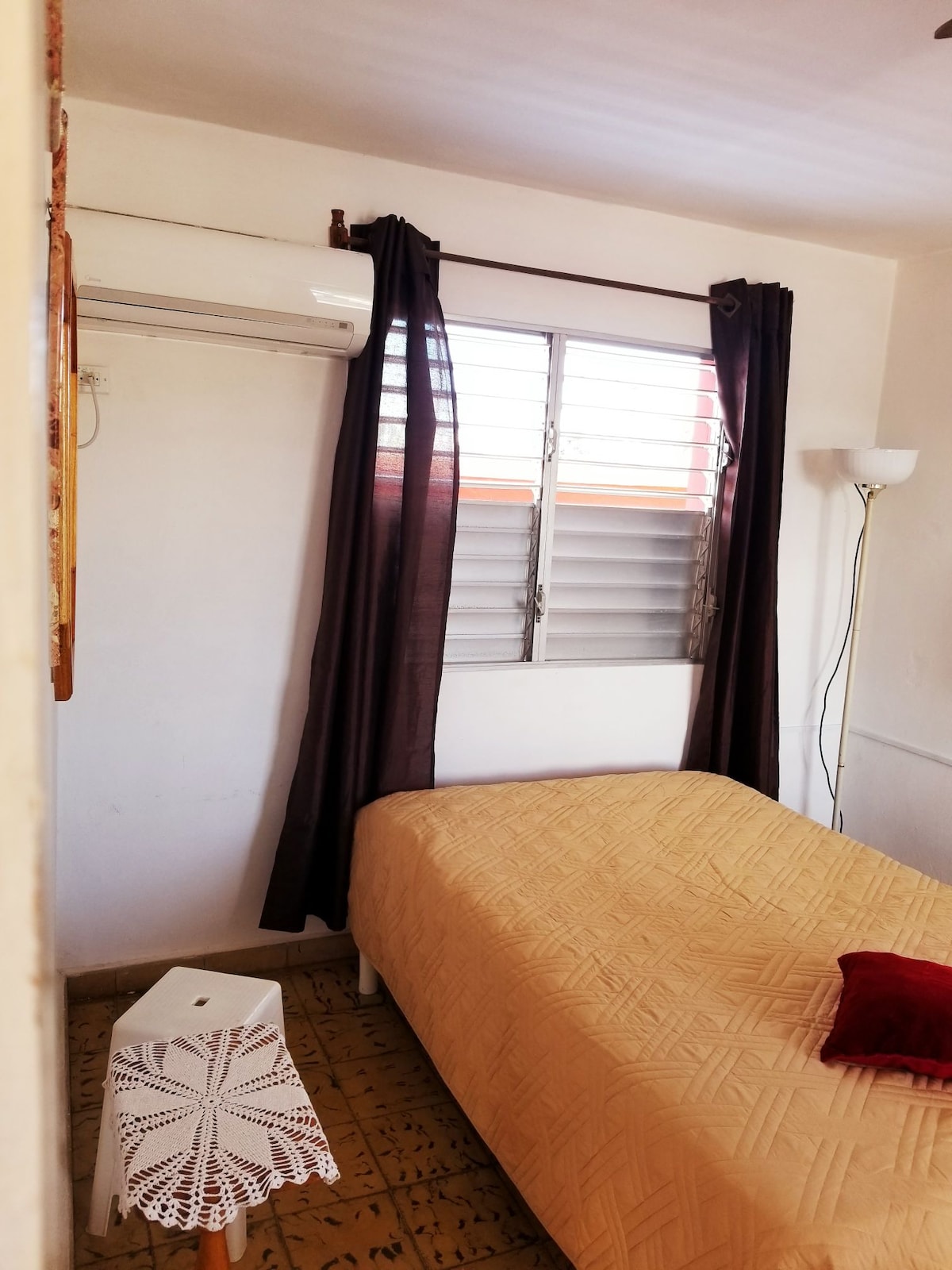 A well-lit sleeping area features a spacious bed covered with a light yellow blanket. A small white side table sits beside the bed. Dark curtains frame a window with two horizontal slats, allowing natural light to enter. An air conditioning unit is mounted above.