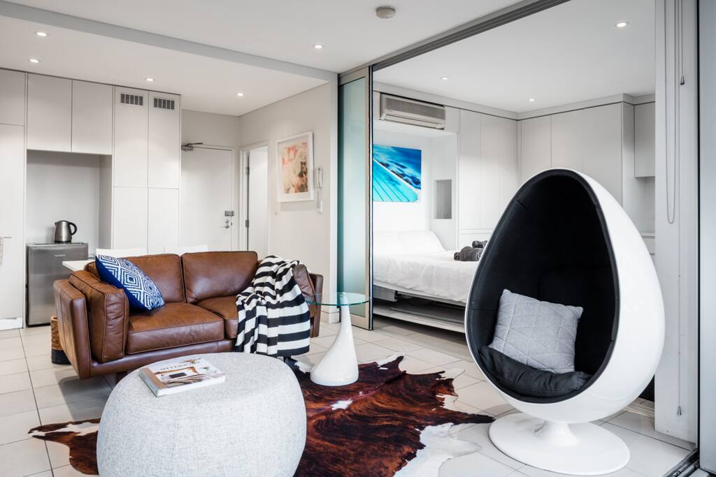 New leather sofa and designer egg chair, cowhide rug, sliding glass panel between living area and bedroom, new Daikin air conditioner (installed since photo taken), table and two chairs behind sofa.