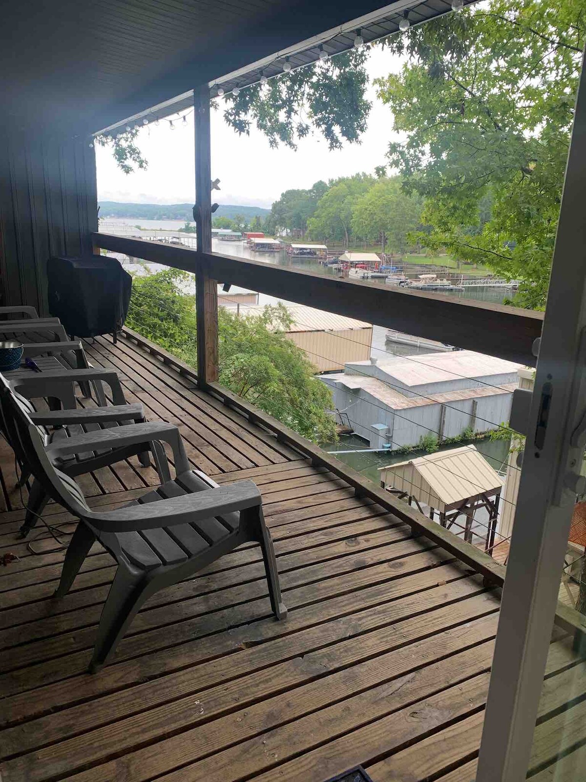 A covered balcony features several gray chairs situated on wooden decking. A railing provides a view of the surroundings, including a glimpse of the lake and nearby marina structures. Lush greenery offers a natural backdrop, enhancing the outdoor space.