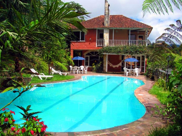 A large natural pool is surrounded by lush greenery, with sun loungers and umbrellas placed nearby. A two-story house with a terracotta roof and balconies overlooks the pool, enhancing the tranquil outdoor space.