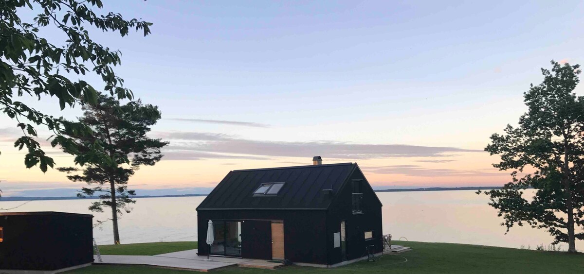 A modern black house is positioned by the serene lake, reflecting the soft hues of the evening sky. Large windows allow for abundant natural light, and a wooden deck extends toward the water, providing a peaceful space for relaxation.