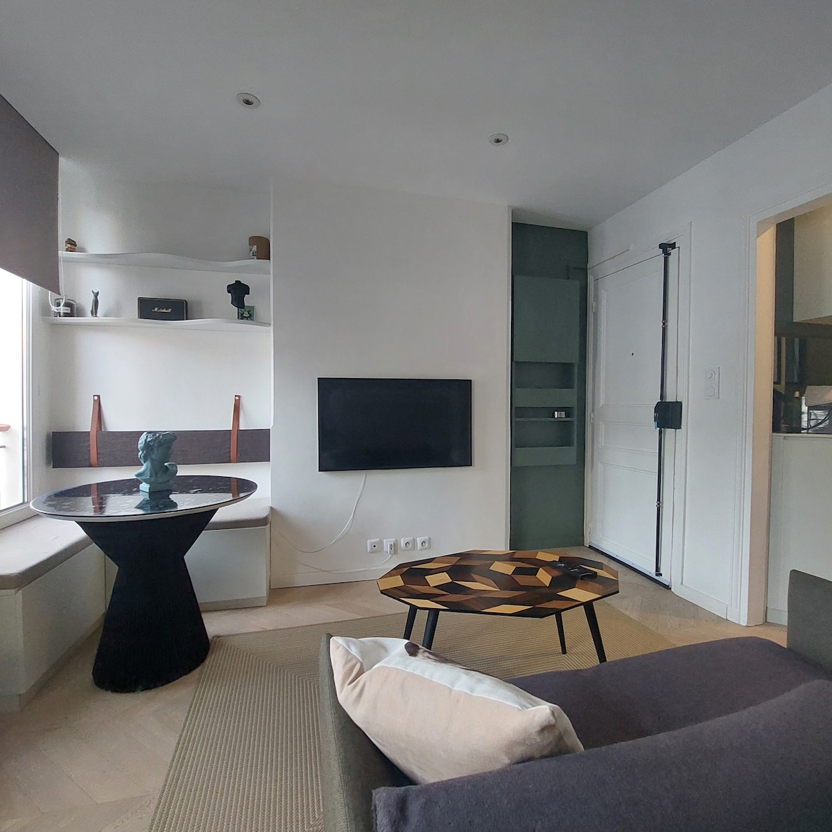 A modern living area features a black round table with decorative elements and a patterned coffee table. A comfortable sofa offers neutral cushions. A television is mounted on the wall, and natural light enters through a nearby window, enhancing the minimalist design.