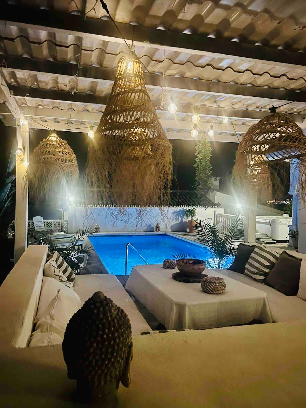 A covered outdoor area features woven pendant lights hanging from a slatted roof. A dining table is set with decorative items, facing a private pool illuminated in blue. Lush plants surround the space, creating a serene environment for relaxation.
