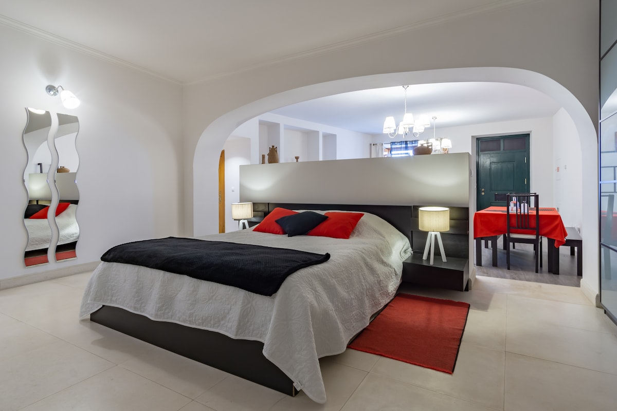 A spacious sleeping area showcases a large bed with a white quilt and layered black and red accents. Nightstands are positioned on either side of the bed, each featuring a lamp. A dining area with a red tablecloth and chairs is visible in the background.