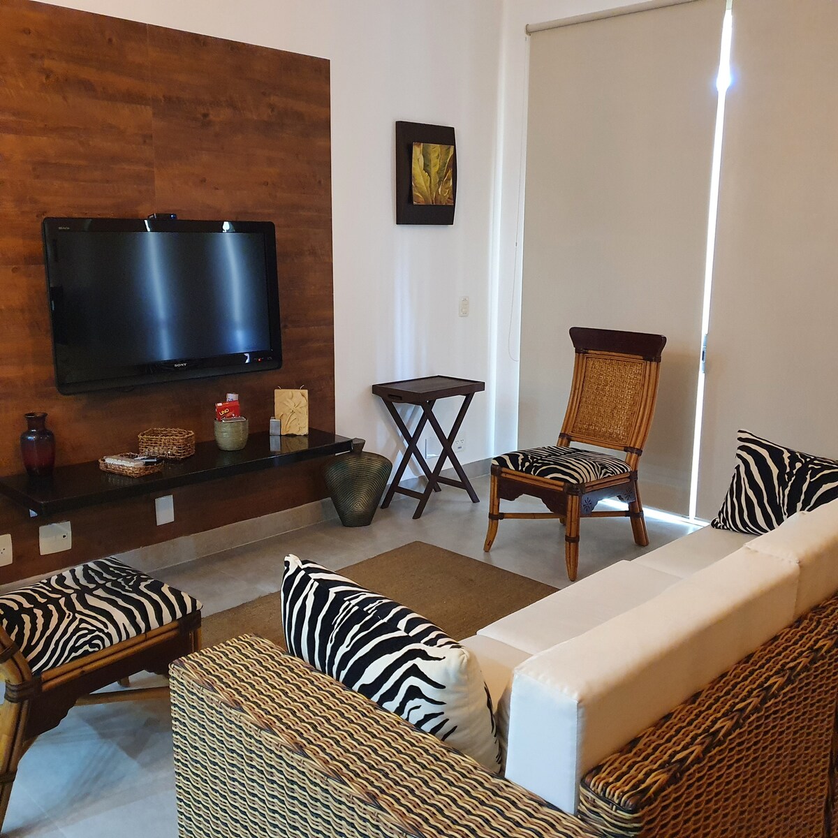 A cozy living area features a large flat-screen television mounted on a wooden accent wall. Comfortable seating includes a white sofa and two woven chairs, each adorned with black and white stripes. Natural light enters through a partially closed window with light-colored blinds.