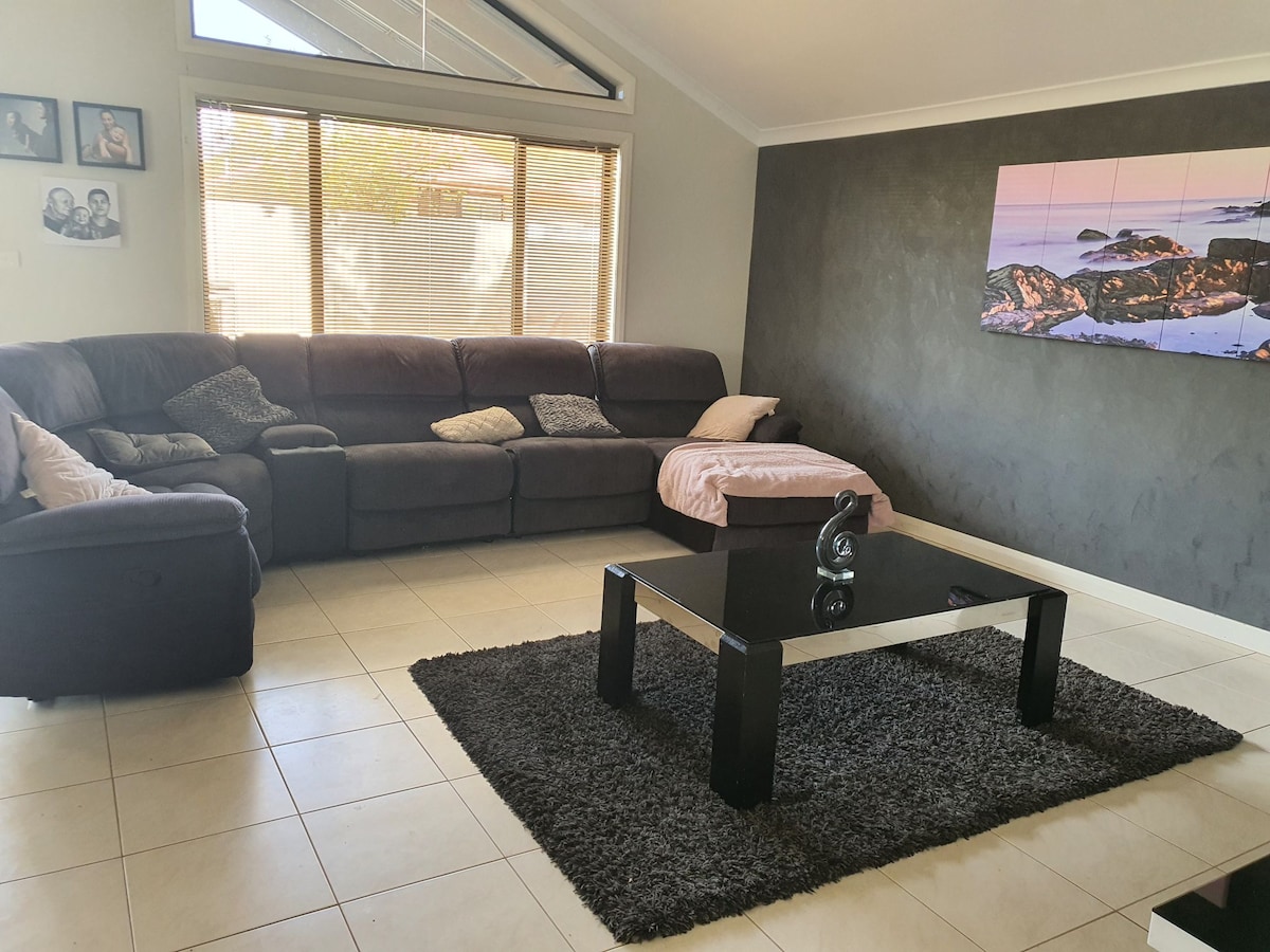 A comfortable living area features a large sectional sofa positioned around a modern coffee table. A cozy rug covers the tiled floor, and natural light floods in through large windows, enhancing the open feel. A framed wall photo display adds a personal touch.