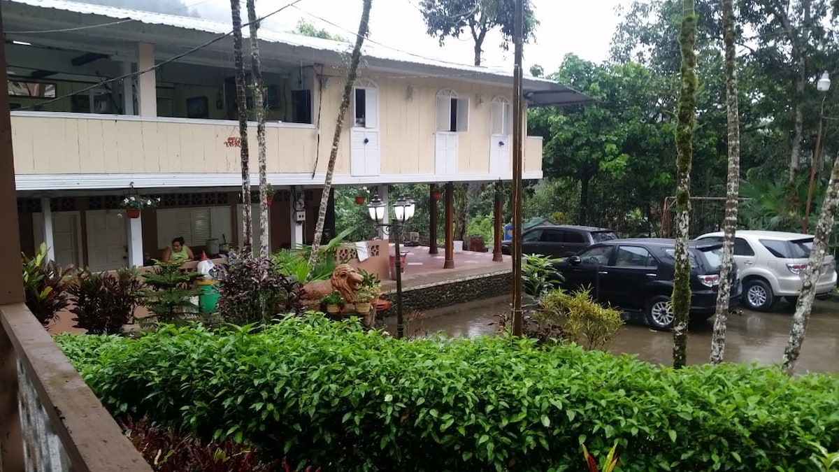 A two-story building is surrounded by lush greenery, with a patio area visible at the front. Several vehicles are parked nearby on the stone pathway, and a small decorative garden features natural elements. The overall setting is tranquil and inviting.