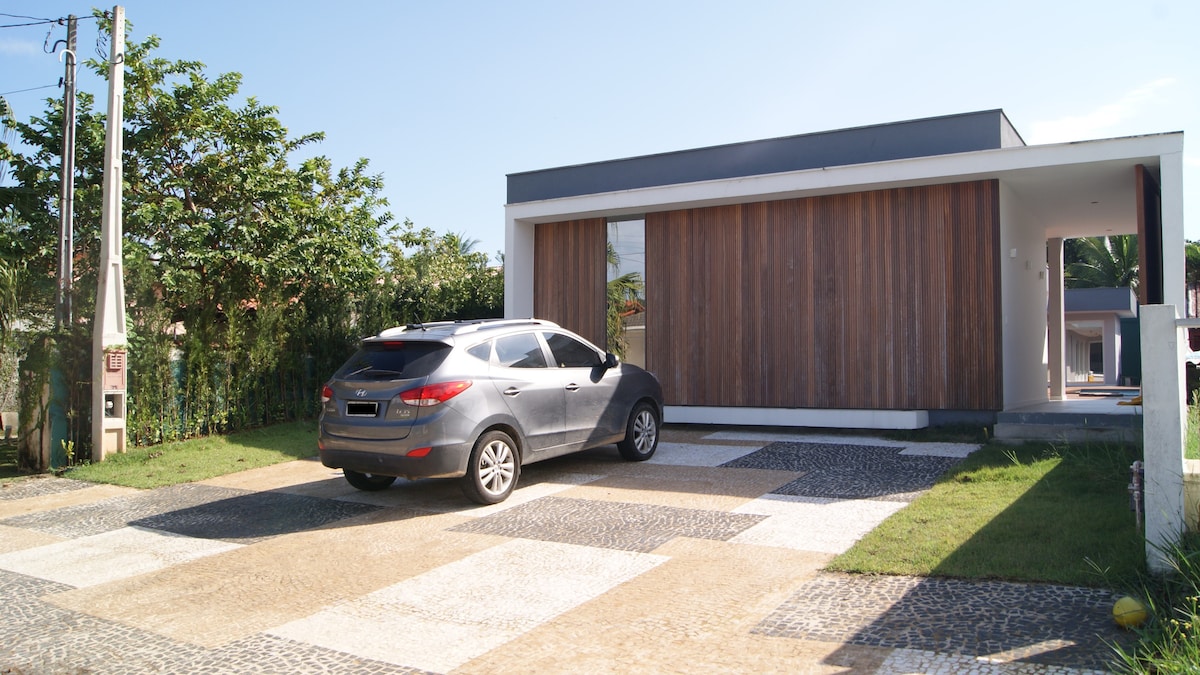 A modern single-story house features a wooden panel door and a spacious driveway paved with stones. A parked car is visible in front, alongside neat grass and a landscaped area with small trees, providing an open and airy exterior.