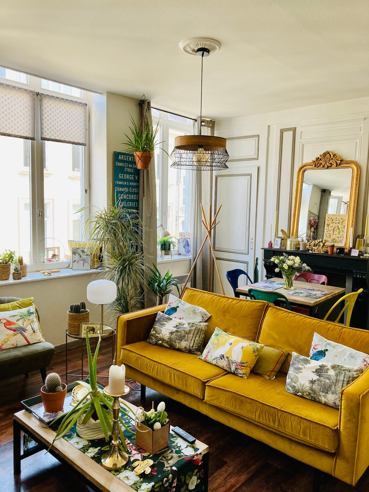 A bright living area features a yellow sofa adorned with patterned cushions. A wooden coffee table holds various plants and decorative items. Large windows allow natural light to fill the space, which is complemented by a vintage-style mirror and a hanging light fixture.