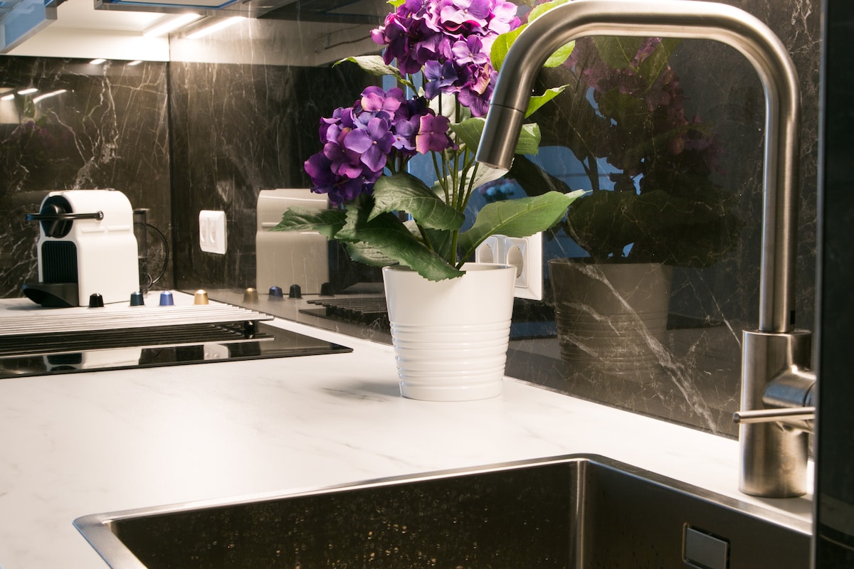 A modern kitchen counter is featured with a sleek stainless steel sink and faucet. A white planter with purple flowers adds a touch of color against the dark marble backdrop. A coffee machine and various kitchen tools are neatly arranged nearby.