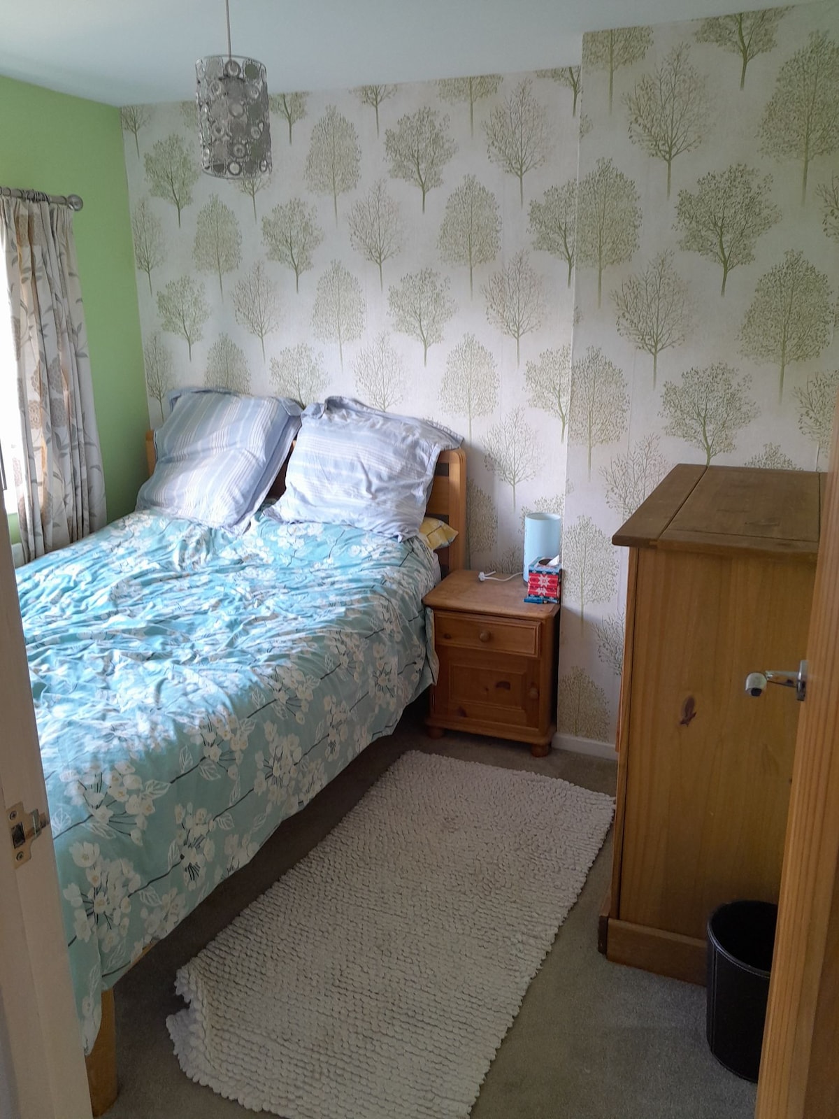 A cozy bedroom is furnished with a spacious bed covered in a floral comforter and decorative pillows. Light-colored wood furniture including a nightstand and wardrobe complements the nature-themed wallpaper. A soft rug is positioned beneath the bed, adding warmth to the space.
