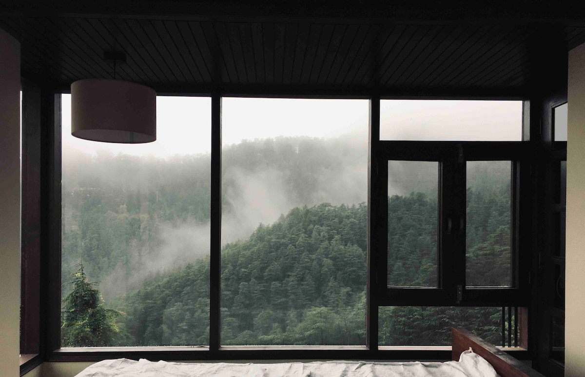A large window provides a panoramic view of mist-covered hills and dense forest, enhancing the connection to nature. Soft morning light filters through the glass, illuminating a cozy sleeping area with a neatly arranged bed frame and white bedding.