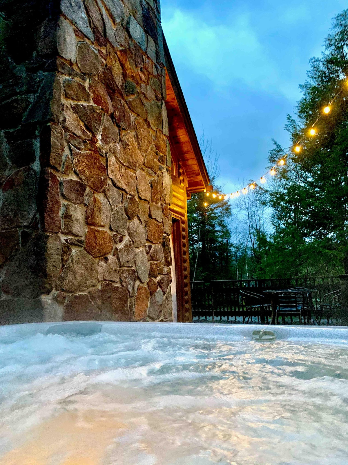 A new hot tub is visible in the foreground, surrounded by stone walls and illuminated by outdoor string lights. The evening sky casts a soft blue hue, while the surrounding greenery creates a serene ambiance.