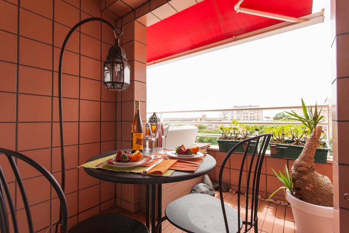 An outdoor balcony is furnished with a small round table and two black chairs. A hanging lantern adds charm, while colorful placemats and plates of fruit create a welcoming dining setup. Lush potted plants line the railing, offering a glimpse of greenery beyond.