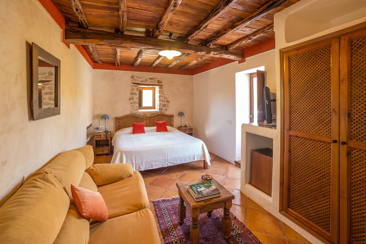 A spacious room is highlighted by a wide bed with white linens. Simple wooden furniture complements the warm earthy tones of the walls. A rustic ceiling adds character, while a small window invites natural light. A cozy sofa and decorative rug enhance the inviting ambiance.