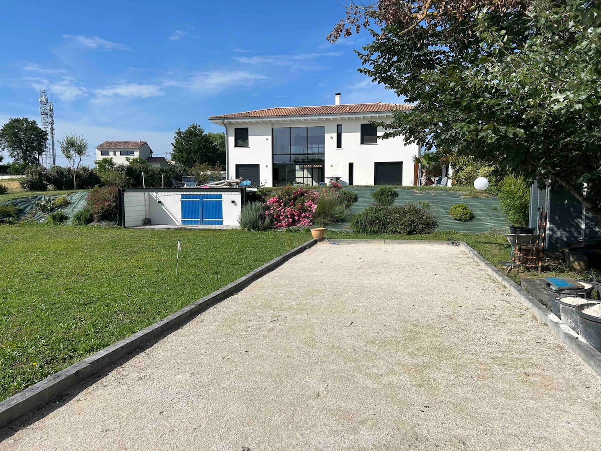 A modern architect-designed house is visible in the background, showcasing large windows on its facade. The foreground features a gravel pétanque court, bordered by a green lawn with shrubs and trees. Clear skies and a well-maintained flower garden enhance the outdoor space.