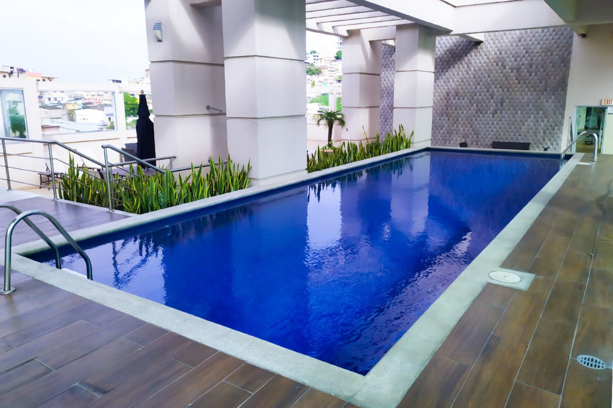 A modern swimming pool is featured with clear blue water and surrounded by sleek tiles. The area is enhanced by greenery along the edges and is complemented by a well-designed structure above, creating a serene environment for relaxation.