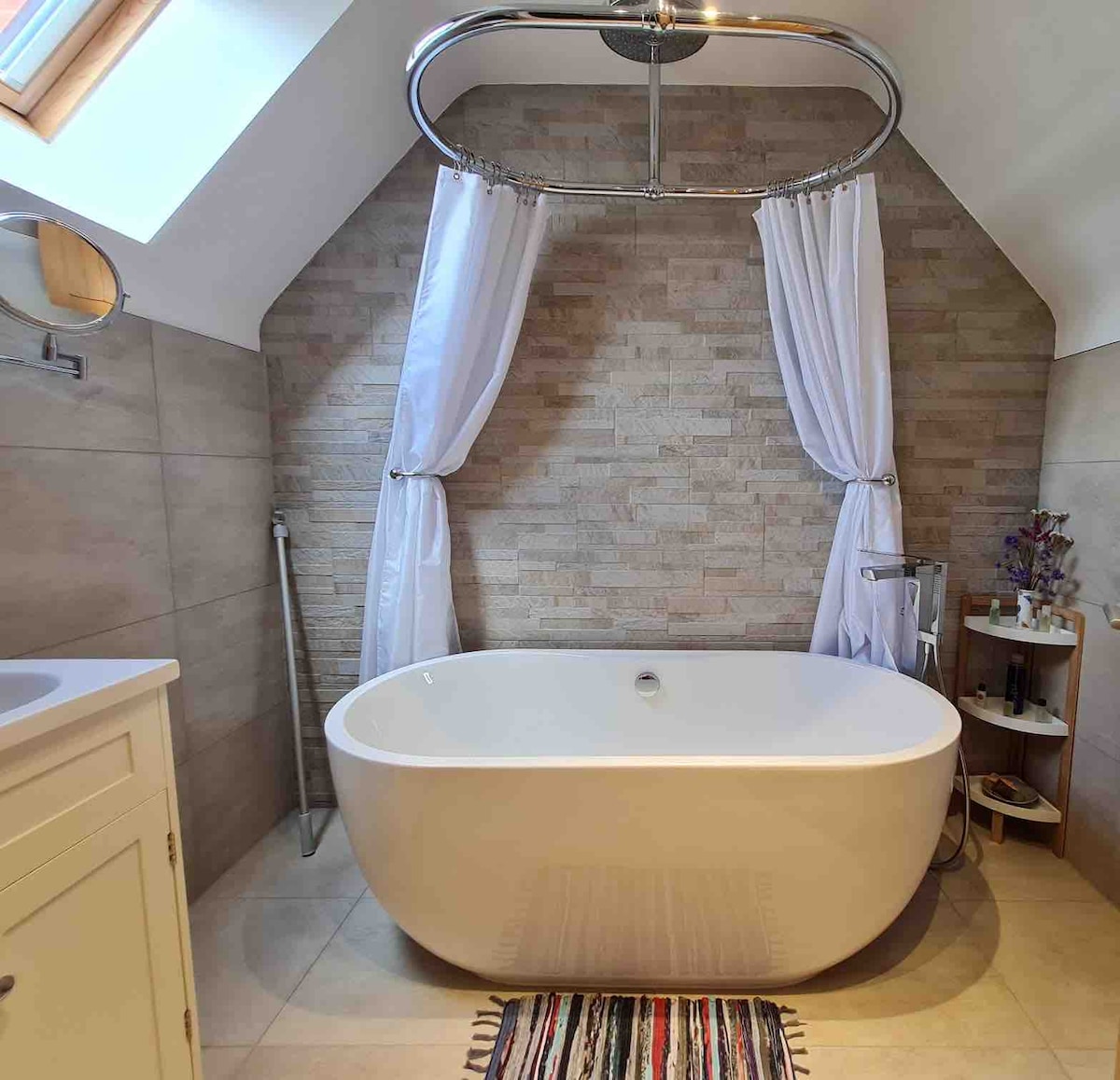 A modern bathroom is showcased with a freestanding bathtub positioned against a textured stone wall. Sheer white curtains frame a skylight above, allowing natural light to enhance the space. A wooden stool with decorative items sits nearby, completing the relaxing ambiance.