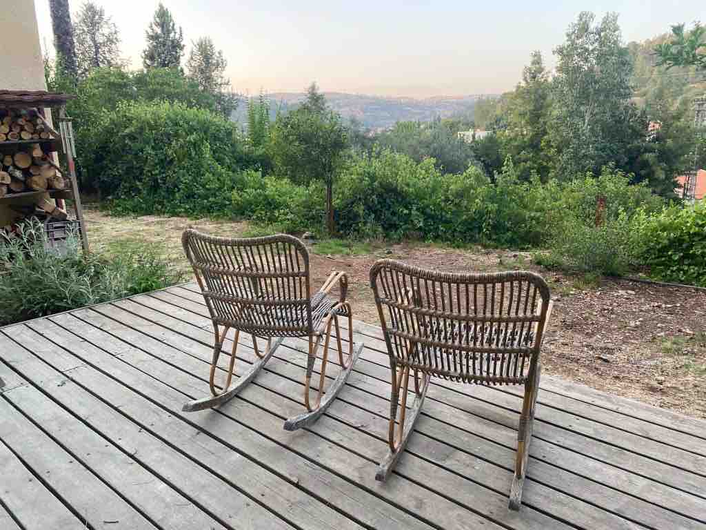 Two rustic rocking chairs made of wicker are positioned on a wooden deck, facing a view of rolling hills and greenery. The surrounding landscape is dotted with trees, creating a sense of tranquility and connection with nature.