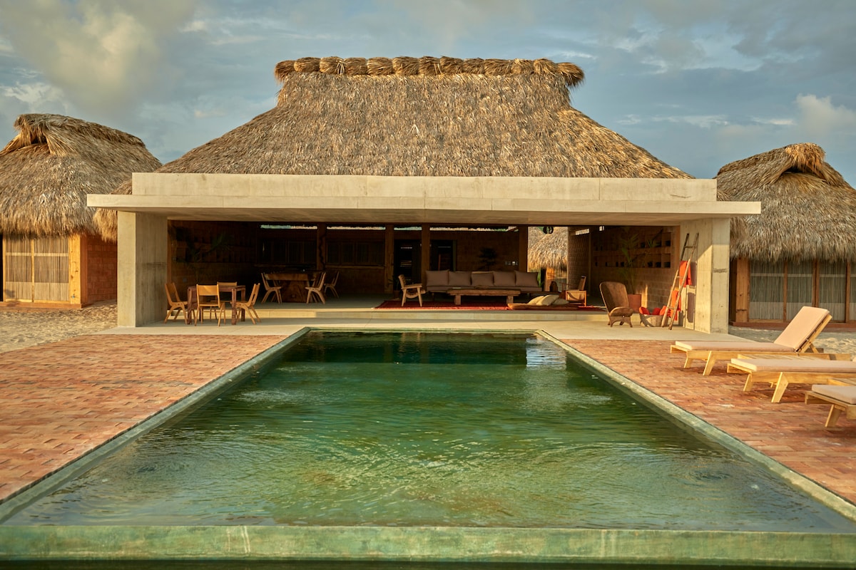 A serene pool reflects the sky and is surrounded by a brick patio. Large thatched roof structures provide shade, with lounge chairs positioned nearby for relaxation. The open-air design allows for unobstructed views of the inviting space and natural surroundings.