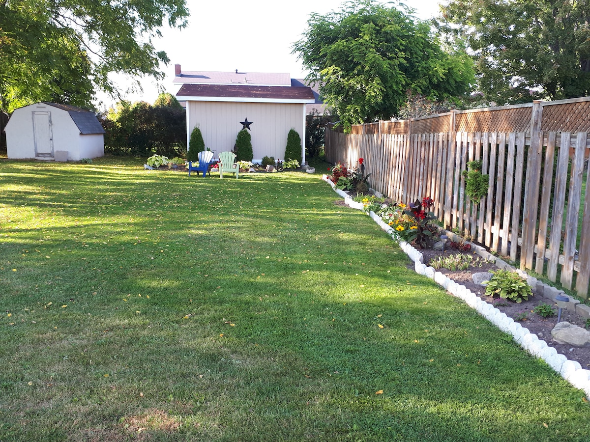 A spacious backyard is presented, featuring a manicured lawn bordered by colorful flower beds along one side. A wooden fence provides privacy, and a small garden shed can be seen in the background. A few chairs are arranged for relaxation in the yard.