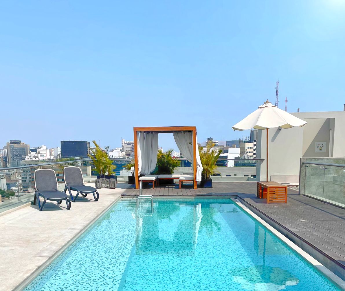 A spacious rooftop area features a refreshing pool surrounded by lounge chairs and a shaded seating area with a pergola. The clear sky and city views provide a serene backdrop, with potted plants adding greenery to the scene.